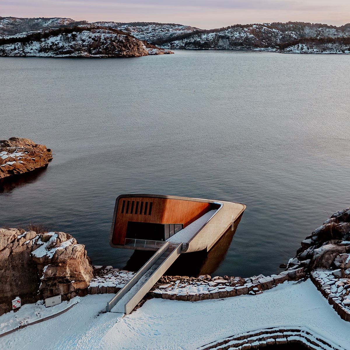 Under 最令人震撼的第一印象，來自其極具存在感的建築本體。這座由挪威國寶級建築團隊 Snøhetta 操刀的建築，全長約34公尺，宛如一塊傾斜滑入海中的混凝土岩體，有一半深埋於海平面以下五米。粗獷、厚實的外觀不僅呼應北歐海岸的原始地貌，更是為了抵禦強烈洋流與惡劣氣候而生。Snøhetta 向來擅長讓建築成為風景的一部分，而 Under 的設計則進一步模糊了「人造」與「自然」的界線。它既像異物，又隨時間被海水、貝類與藻類慢慢覆寫，成為海洋生態的一環。這並非炫技，而是一種謙遜地向自然低頭的設計態度。