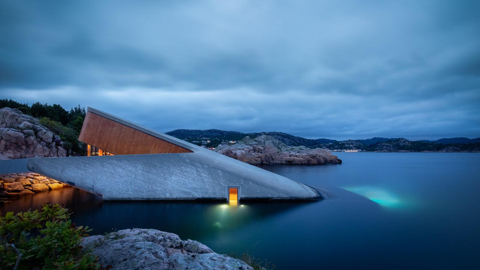 Under 最令人震撼的第一印象，來自其極具存在感的建築本體。這座由挪威國寶級建築團隊 Snøhetta 操刀的建築，全長約34公尺，宛如一塊傾斜滑入海中的混凝土岩體，有一半深埋於海平面以下五米。粗獷、厚實的外觀不僅呼應北歐海岸的原始地貌，更是為了抵禦強烈洋流與惡劣氣候而生。Snøhetta 向來擅長讓建築成為風景的一部分，而 Under 的設計則進一步模糊了「人造」與「自然」的界線。它既像異物，又隨時間被海水、貝類與藻類慢慢覆寫，成為海洋生態的一環。這並非炫技，而是一種謙遜地向自然低頭的設計態度。