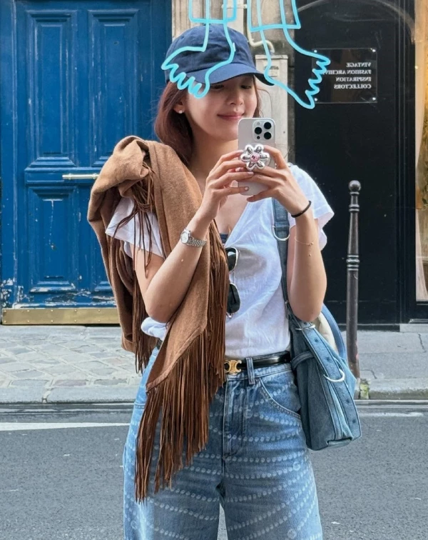 這張穿搭照，展現了章若楠非常隨興層次感的一面！比起上一套的活潑撞色，這身造型選用了大面積的藍色丹寧褲與白T-shirt作為搭配基調，瞬間帶出一種很美式率性的時髦氛圍。最吸引我目光的，便是她隨意披在肩上的那件大地色流蘇披肩，在巴黎街頭的背景下，更添波希米亞式的浪漫，並也與深藍色棒球帽，形成了一種完美「深淺對比」的混搭感，讓簡單的穿搭，瞬間有了視覺重點。完全就是一般人也能輕鬆效法的質感私服搭法！