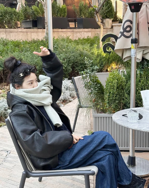 我一直覺得，章若楠的私服最迷人之處，不在於堆疊了多少當季爆款，而是在於那股恰到好處的鬆弛感。看看這張在露天咖啡座的穿搭照，她選擇了一件極具存在感的寬廓形皮外套，那種皮革特有的冷冽光澤，被頸間厚實的淺灰織紋圍巾完美中和了，剛柔並濟。最讓人有感的便是那高高紮起的丸子頭，以及隨手推在髮際線上的墨鏡。這樣看似隨意的小動作，卻能在厚重的層次穿搭中，更加透出一種「Well〜我只是隨便穿穿」的美式輕鬆感。不妨多加參考吧。