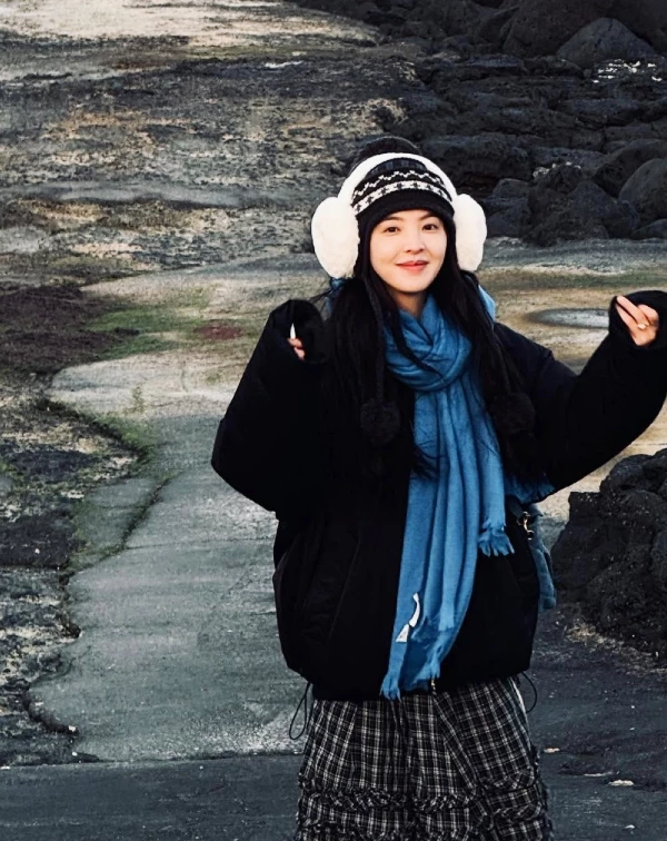 面對冷風颼颼的海景，章若楠選用全黑的廓型羽絨外殼作為戶外裝扮，展現出她獨有的率性感。最吸睛的亮點，在於她聰明地運用了天藍色長圍巾進行跳色點綴，在深色背景前顯得格外活潑搶眼；頭上那頂帶有黑白圖騰的針織帽，加上兩側超大的白色毛絨耳罩，更是可愛與實用的終極結合，在暖陽映照下不僅襯托出鄰家女孩般的親切朝氣，更賦予整體造型一種精緻的「大人可愛」質感。