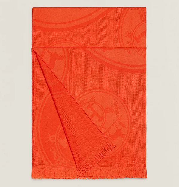 這條Hermès圍巾上不僅飾有經典駿馬Logo緹花刺繡，更以耀眼的愛馬仕橘點綴，為秋冬沉寂的造型注入亮眼焦點。